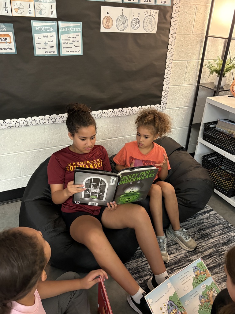 Two children sit on a bean bag chair and read books. Another child stands nearby. A bulletin board with paper is behind them.