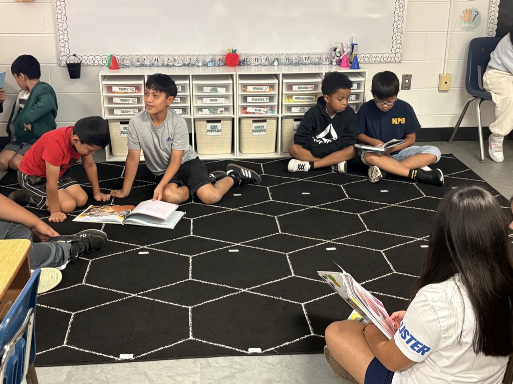 Several children sit on a rug in a classroom. They hold books and read. A teacher is seated among them.