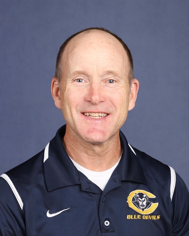 A man smiles at the camera, wearing a blue polo shirt with "Blue Devils" text and a yellow logo.