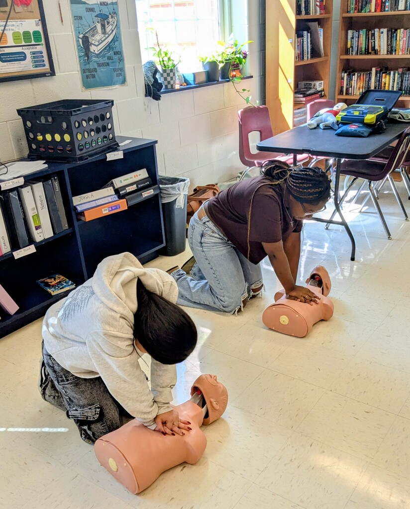 Two people practice CPR on manikins in a classroom with bookshelves, a table, and chairs.