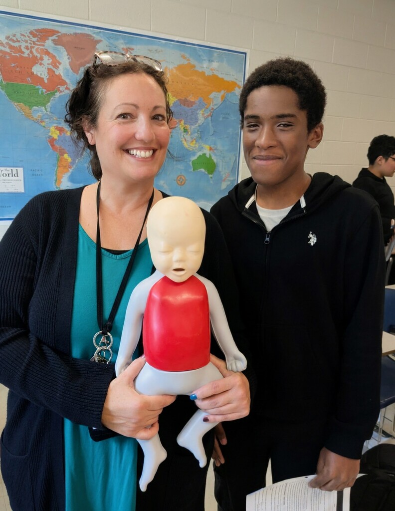 Woman holding a CPR mannequin in class. Another student with a paper stands next to her.