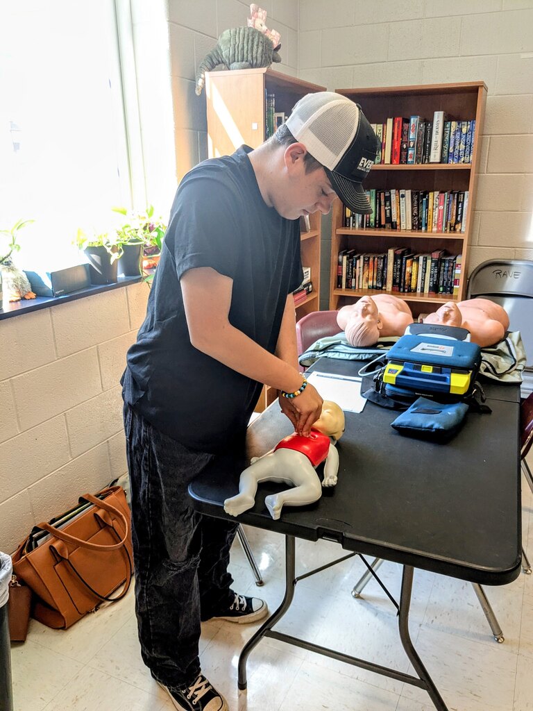 Man in a black shirt and baseball cap performs CPR on a manikin on a table.