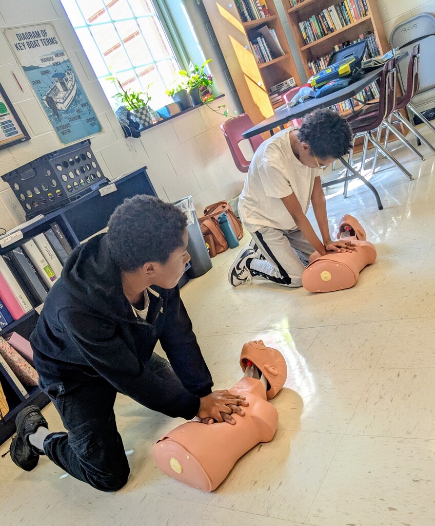Two students practice CPR techniques on manikins in a classroom with bookshelves and chairs.
