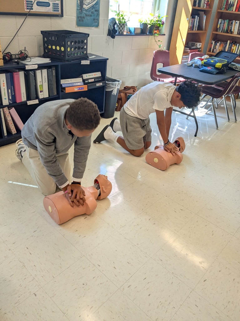 Two students practice CPR on mannequins. One kneels, the other on hands and knees. Books and tables in background.