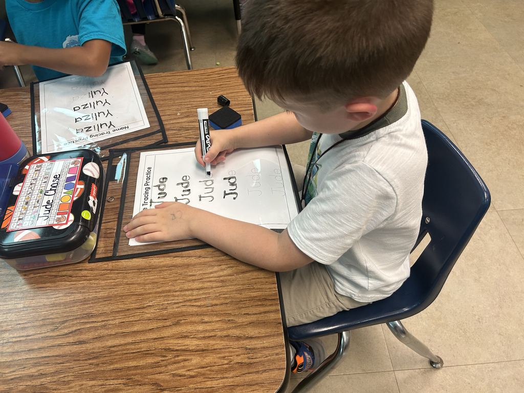 A young boy in a white shirt is writing on a white sheet of paper at a desk.