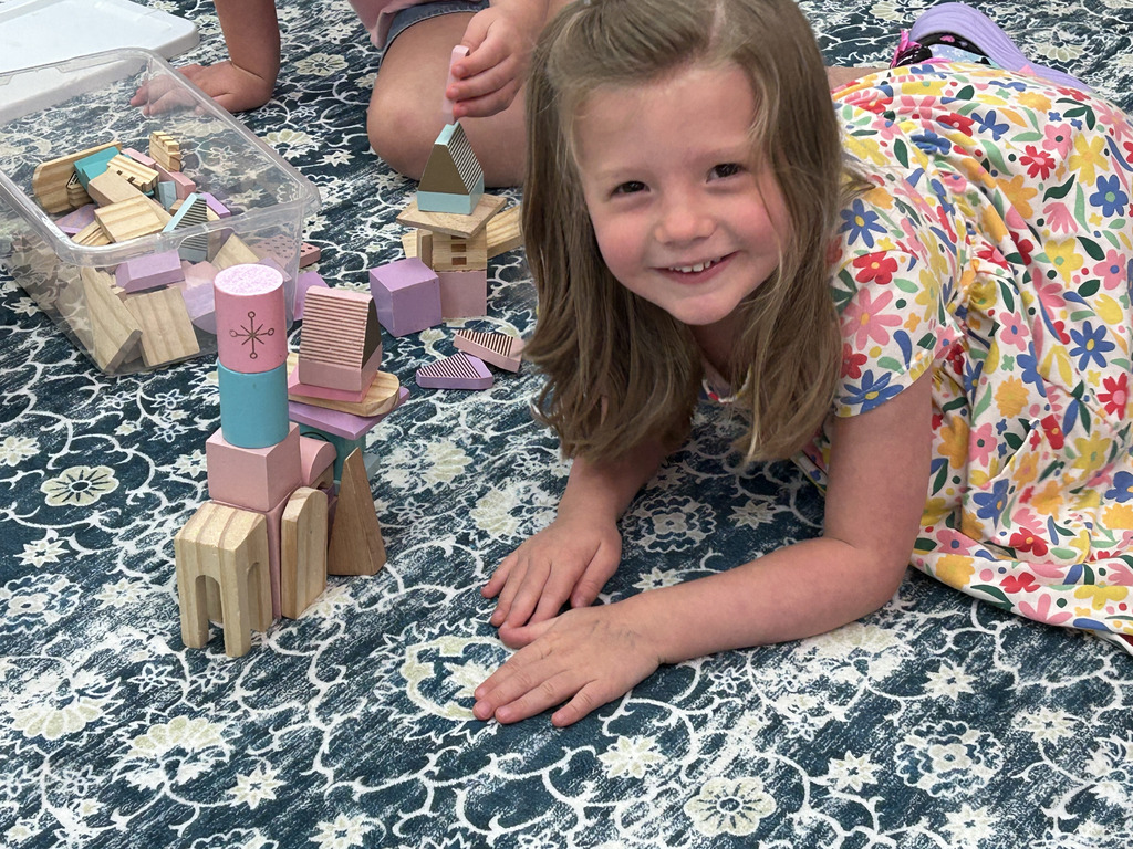 A child lies on a patterned floor, smiling, next to blocks. Other children and toys are nearby.