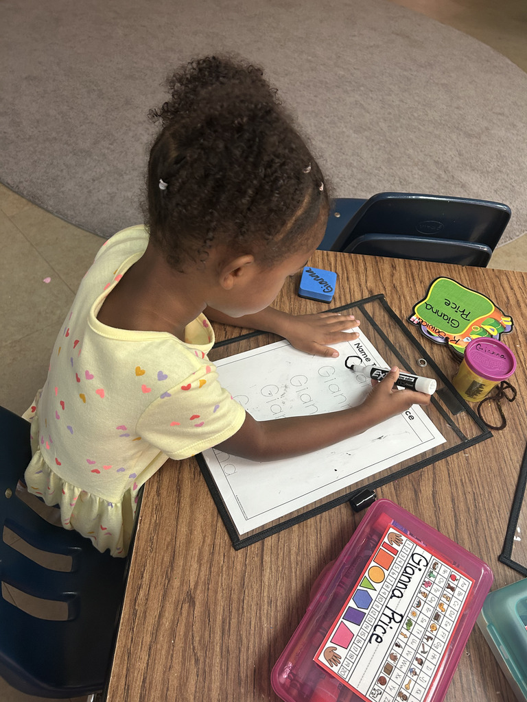 A young girl at a table, seated, using a marker on a dry-erase board with papers and tools.