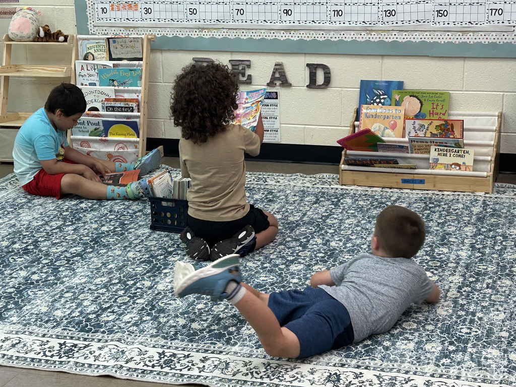 Three children sit on a rug in a classroom. One reads a book, another holds a book, and one lies on the floor.