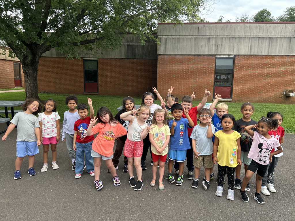 A group of children standing outside near a tree and a brick building, posing with hands raised.