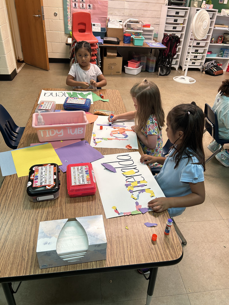 Four children seated at a table, engaging in a creative activity. They use scissors and paper to form letters.