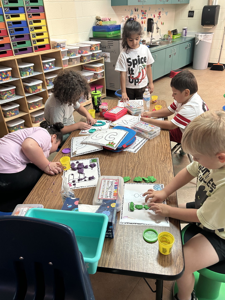 Kids sit around a table working on a project, one uses a stamp. An adult helps. Background has shelves.