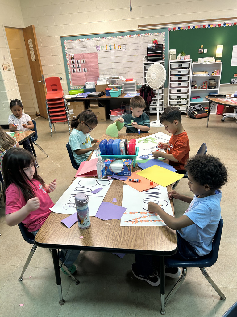 Children sitting at a table, working on a paper craft project. Background includes chairs, tables, and a bulletin board.