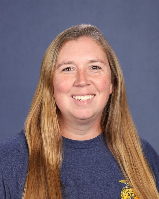 A woman with long blonde hair smiling, wearing a blue t-shirt with a logo on it.