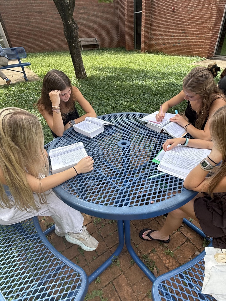 Four individuals are seated at a blue table, each holding open books and writing. Brick building and grassy area in the background.
