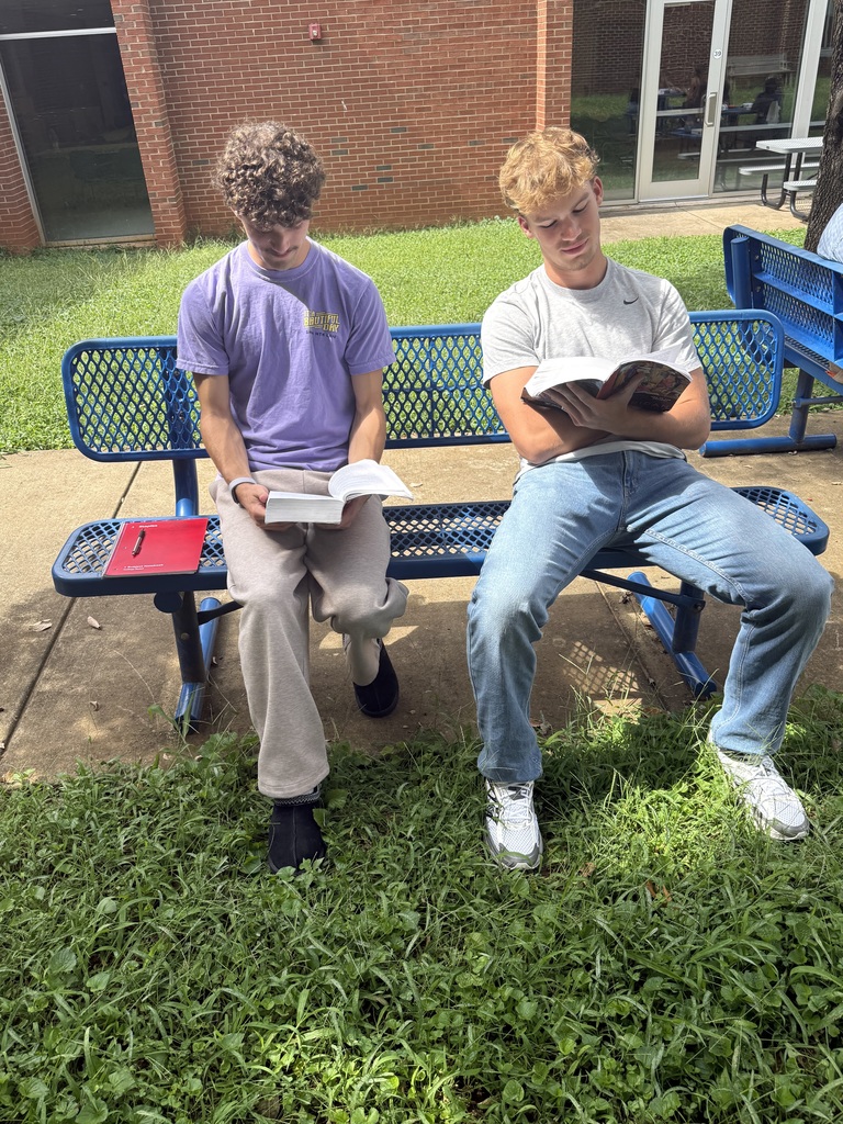 Two young men sit on a blue bench, one reading a book and the other holding a book.