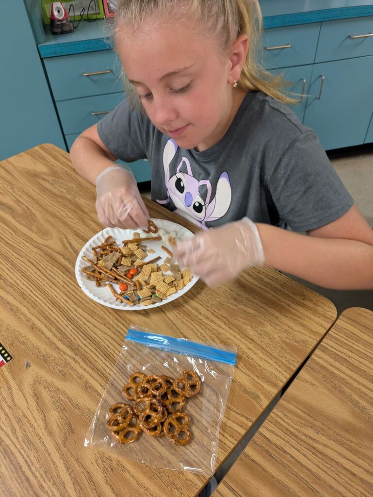 A young girl eats snacks from a paper plate while seated at a school desk.
