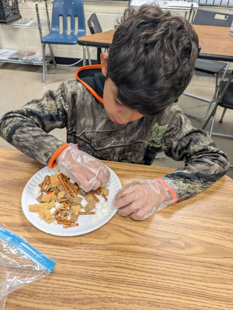 A child with gloves on sits at a table, examining a plate of food. A plastic bag is on the table.