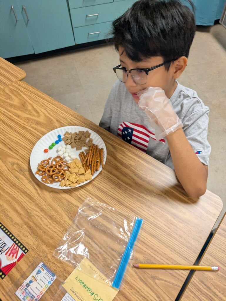 A child in a gray shirt and glasses sits at a table eating from a paper plate.