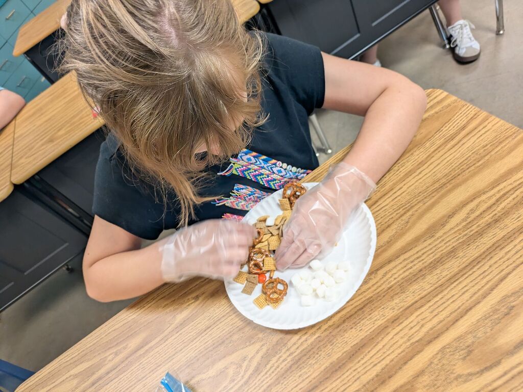 A young child wearing gloves seated at a table, preparing a plate of food. Background shows a classroom.