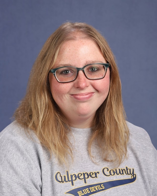 Woman with blonde hair wearing glasses. She is wearing a blue t-shirt with white lettering.