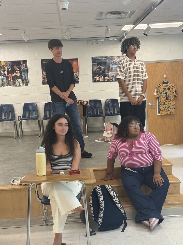 Five people in a classroom. One sits at a desk, another sits on the stairs, and three stand behind them.