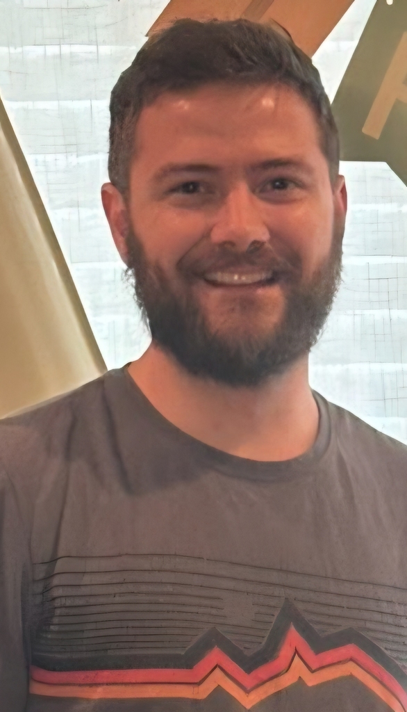 Man smiling with short hair and beard. He wears a gray t-shirt with a mountain design.