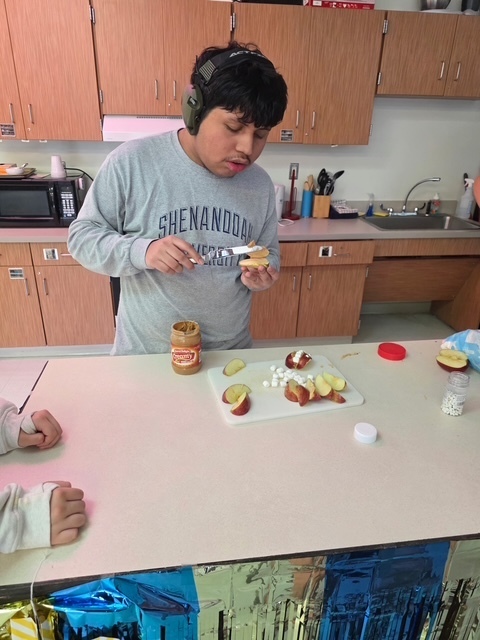 Person in gray shirt eats food in a kitchen. A countertop has peanut butter, sliced fruit, and small containers.