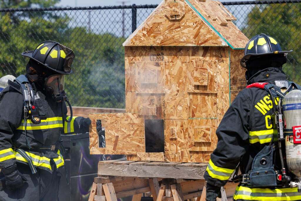 Two CTEC Firefighter instructors in full protective gear and SCBA demonstrate fire behavior using a miniature wooden structure during a hands-on fire dynamics lesson.