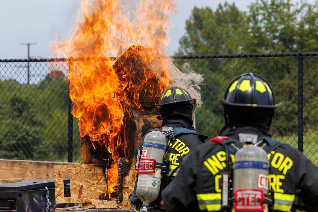 Two CTEC Firefighter instructors in full protective gear and SCBA observe a miniature wooden structure engulfed in flames during a fire dynamics demonstration.