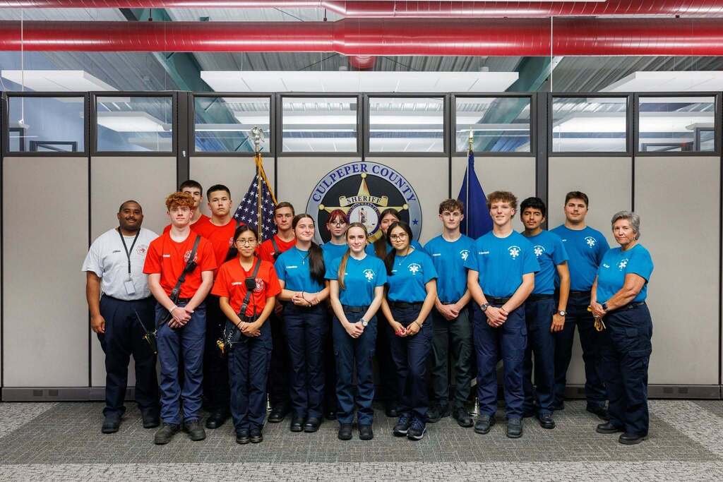 TEC EMT and Firefighter students pose with their instructors during a visit to the Culpeper County Sheriff’s Office. Students wear blue EMT and red Firefighter program shirts and stand in front of the Culpeper County Sheriff emblem and flags.