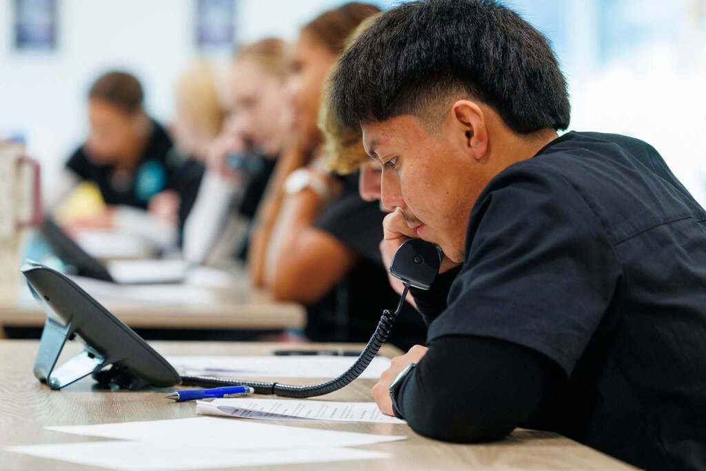A CTEC Medical Assistant student practices professional telephone communication while using a classroom phone and following written materials during the activity.
