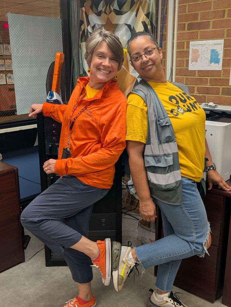 Two women pose, one in orange and one in yellow, both wearing sneakers, in an office.