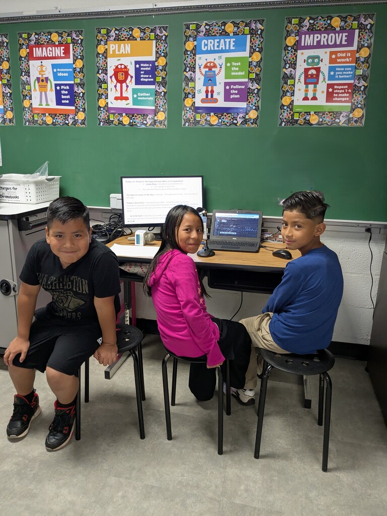 Three children in front of a computer station in a classroom. One uses a laptop, while the others use a desktop.