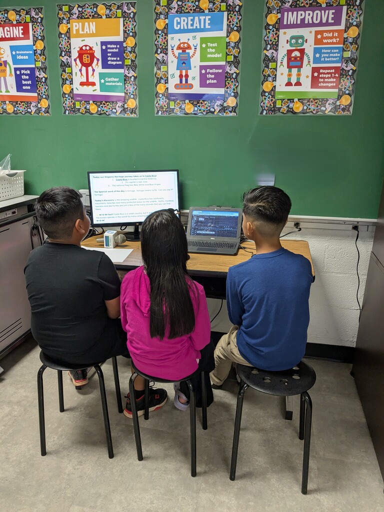 Three students sit at a desk, facing a computer. A green wall displays posters with words like Plan, Create, and Improve.