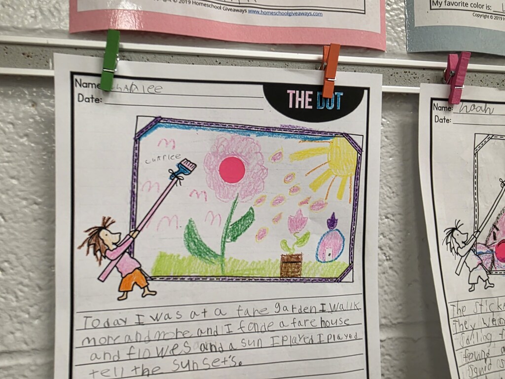 White wall with papers displaying a child's drawing. One paper shows a child holding a rake, and a flower with the words "Today I was at a fair."