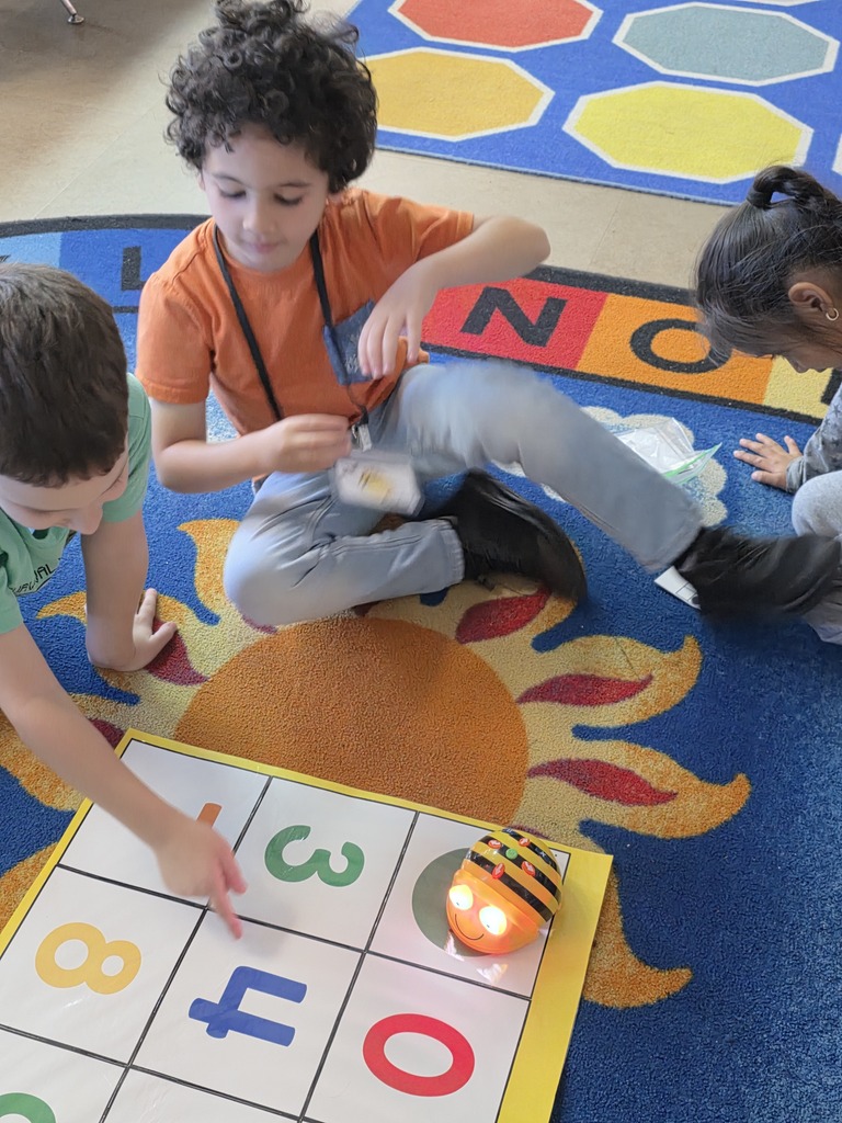 Children sit on a rug with a game board. One child points to letters while another holds a robot.