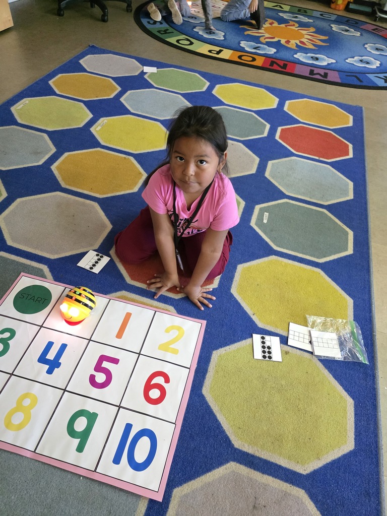 A child in a pink top sits on a hexagonal rug, working on a number grid that ranges from 1 to 10.