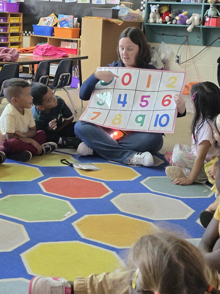 Woman sitting on floor with children holding a number card. Children in chairs on multicolored rug.