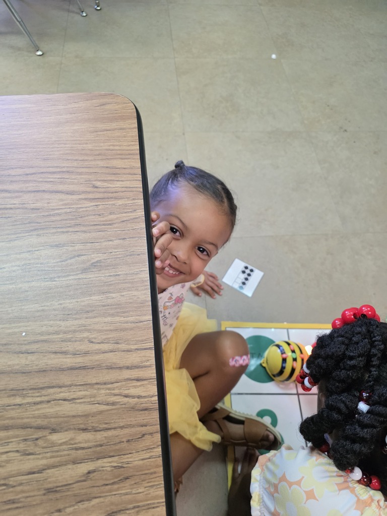 A young child in a yellow dress sits at a wooden table, peeking over it. Another child, in a white dress, sits behind the table.