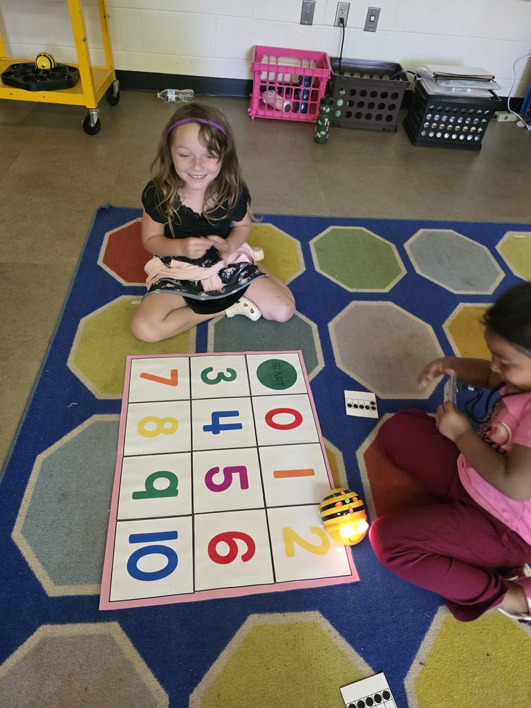 Two children are seated on a multicolored floor mat, engaging in a game with numbered tiles and a small toy.