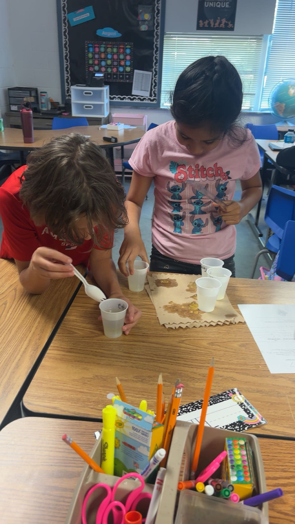 Two children engaged in an activity with cups and materials on a table, with a classroom background featuring desks and a bulletin board.