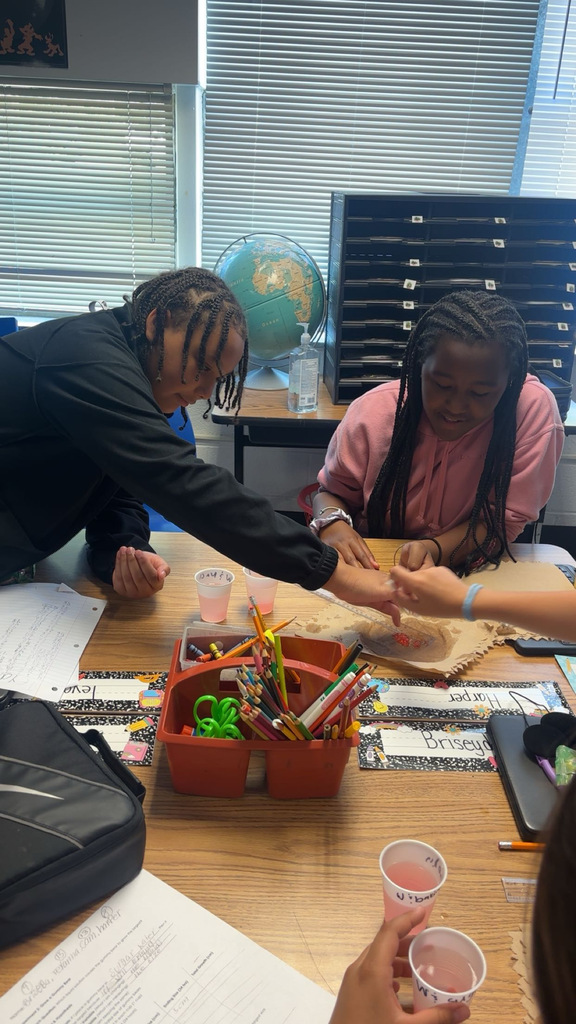 Two students sit at a desk, engaging in a craft activity with pens and scissors. One student reaches over to hand something to the other.