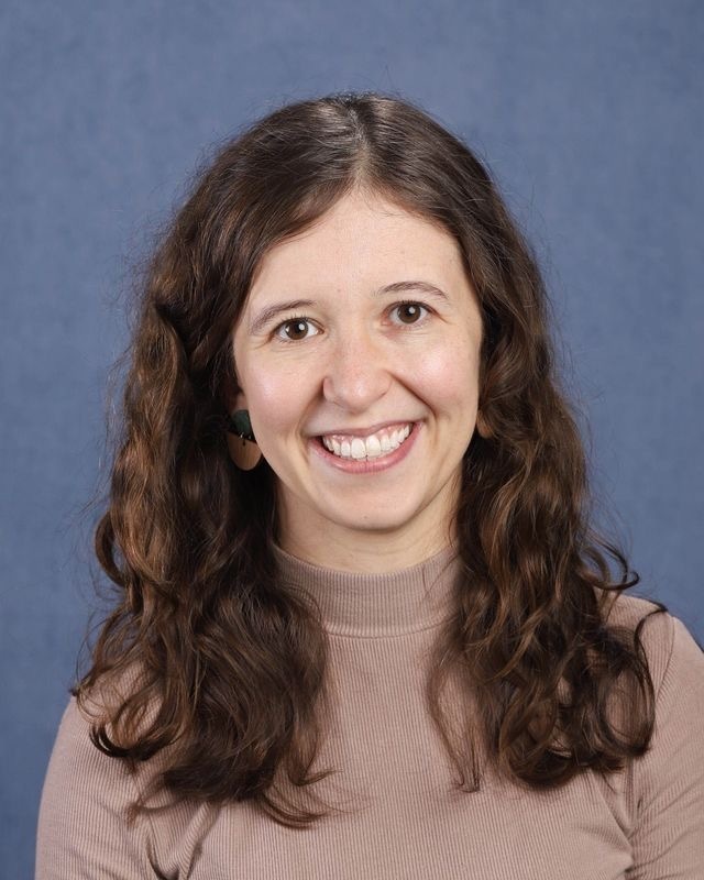 Woman with brown hair, smiling, wearing a beige turtleneck. She has an earring. The background is solid blue.