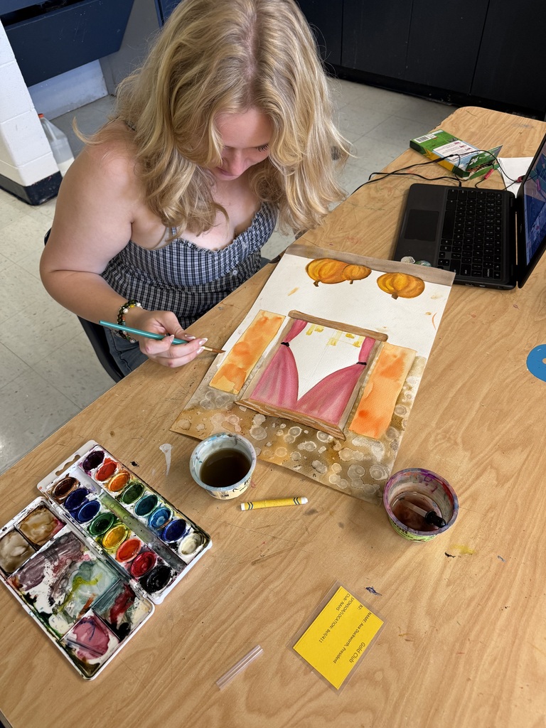 A person with blonde hair sits at a desk, painting a picture on a canvas. A laptop is nearby.