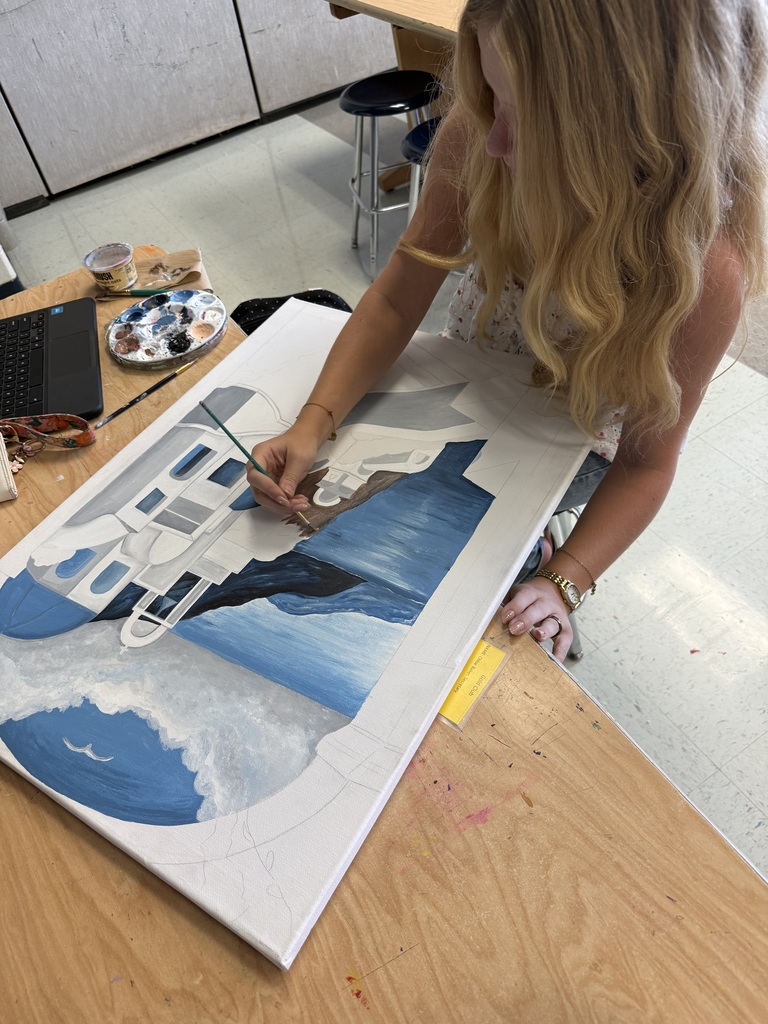 A person painting a picture of a white building on a large blue background. A laptop is nearby.