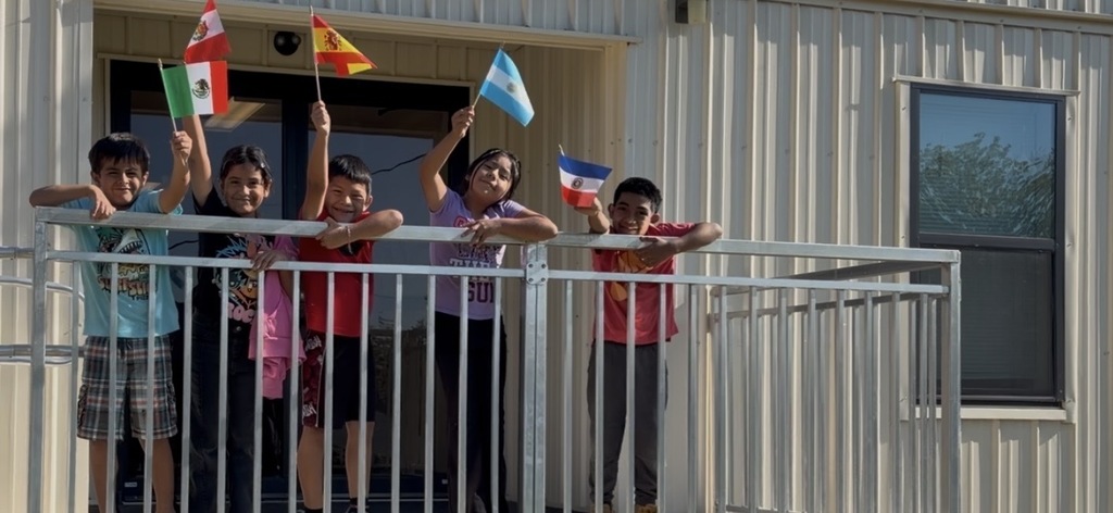 Children holding flags standing on a balcony of a building. The railing is white, and the background is white with windows.