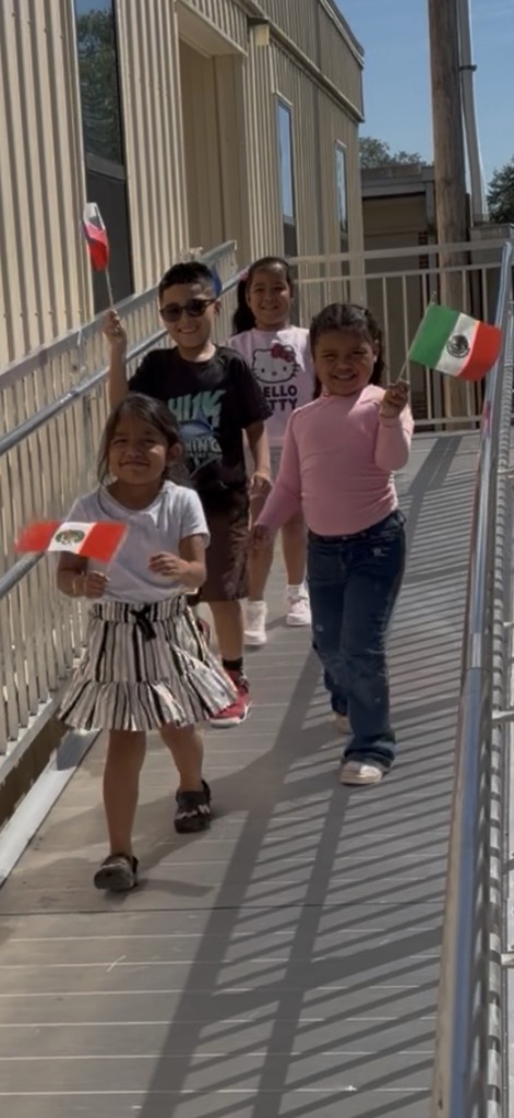 Four children walk on a walkway, holding flags. Two children wear sunglasses. The children are in front of a building with a metal railing.