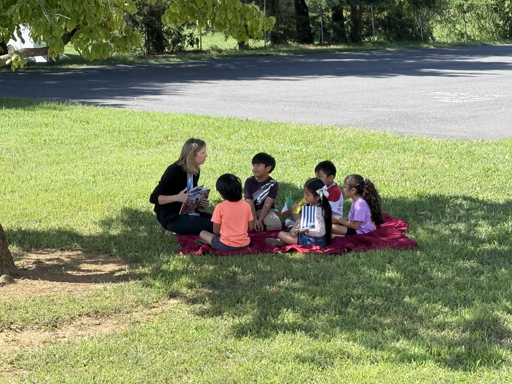 A woman sits on a blanket with several children, reading a book to them in a grassy area.