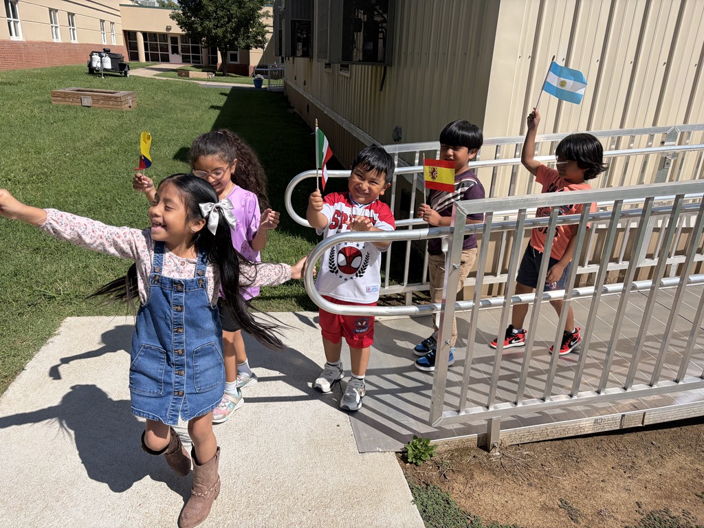 Several children wave flags, walking up a ramp to a building with a grassy area and trees.