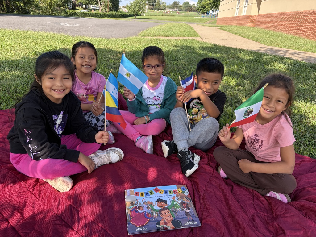 Five young children hold flags, sitting on a red blanket in a grassy area, with a book in the foreground.